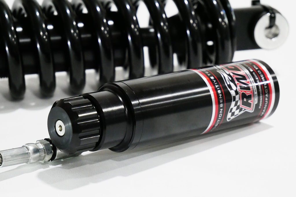 A close-up side shot of an RWD V-Twin RS-2 reservoir, featuring a black body with red and white RWD branding, connected to a silver hose, with a black spring in the background.