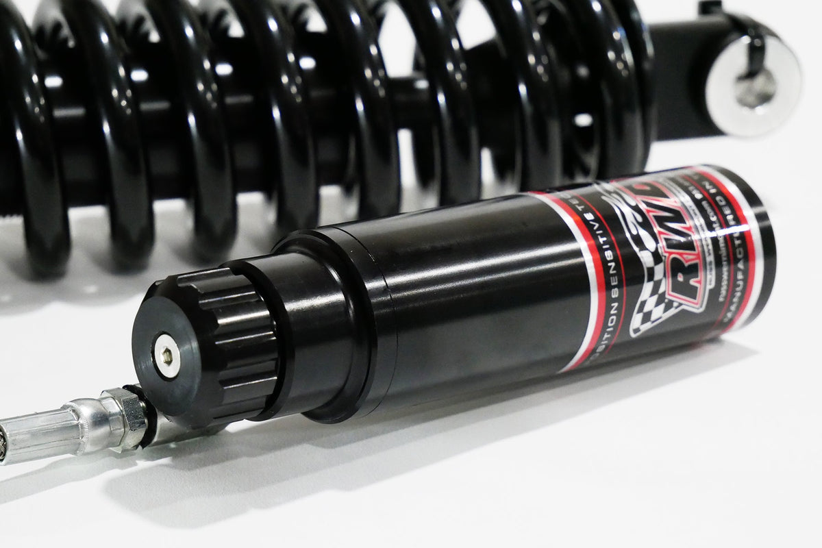 A close-up side shot of an RWD V-Twin RS-2 reservoir, featuring a black body with red and white RWD branding, connected to a silver hose, with a black spring in the background.