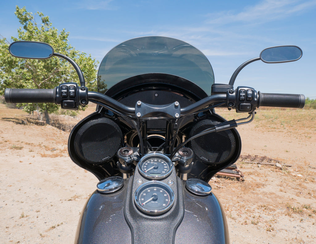 A close-up, eye-level view of the front of a dark gray RWD V-Twin motorcycle, featuring a black fairing with integrated speakers, a clear windshield, black handlebars with mirrors, and chrome gauges on the fuel tank. The motorcycle is parked outdoors on a dirt path with a tree and blue sky in the background.